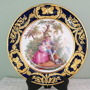 May include: A round decorative plate featuring a central illustration of two figures in period clothing with a birdcage, surrounded by a deep blue border with ornate gold leaf butterfly motifs and intricate scrolling foliage patterns.