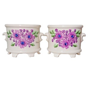 May include: Two off-white ceramic planters with side handles and four small feet. Each planter features a hand-painted floral design with purple daisy-like flowers, blue centres, and green leaves. The left planter has small text reading Rose Song.