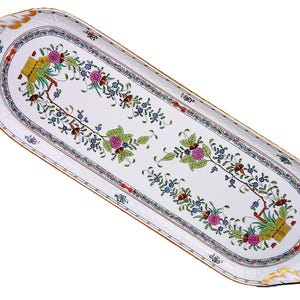 Puede incluir: Una bandeja ovalada de cerámica blanca con un diseño floral. La bandeja tiene un borde dorado y está decorada con flores y follaje rosas, azules y verdes. El diseño es simétrico, con patrones florales.