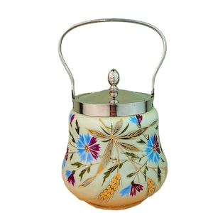 Op de afbeelding: Een crèmekleurige glazen koekjestrommel met een zilverkleurig metalen handvat en deksel. De pot is voorzien van een handgeschilderd bloemmotief met blauwe en paarse bloemen, groene bladeren en gouden korenaren. Het deksel heeft een kleine zilverkleurige knop.