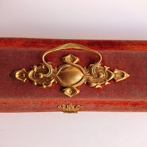 May include: A rectangular storage box covered in red fabric with a prominent gold-toned metal handle featuring an ornate, symmetrical design. The box includes a small, decorative gold-toned metal latch centered along the bottom edge.