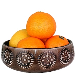 May include: A round, brown ceramic bowl featuring a repeating embossed sunburst pattern with small dots. The bowl is filled with a collection of fresh citrus fruits, including one yellow lemon and three bright orange oranges.