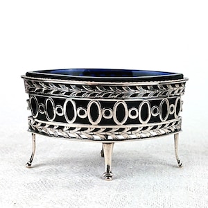 Può includere: Una saliera antica argentata con un inserto in vetro blu cobalto. Il contenitore ovale presenta un design traforato decorativo con motivi ovali e a foglie. Poggia su quattro piccole gambe curve.