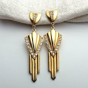 May include: A pair of Art Deco-style gold-tone earrings. Each earring features a fan-shaped top with a row of clear rhinestones, a central geometric design, and a dangling, vertical, ridged bottom. The earrings are displayed against a white background.