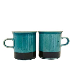May include: Two ceramic mugs featuring a two-tone design with a speckled teal upper section and a solid black base. Each mug has a cylindrical body, a smooth rim, and a curved handle on the side, showcasing a consistent colour scheme and finish.