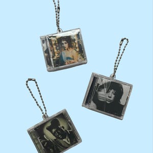 Puede incluir: Tres llaveros de plástico transparente con forma de estuche de CD con imágenes en blanco y negro en el interior. Cada llavero tiene una cadena de bolas plateada. Las imágenes incluyen un retrato vintage y otros dos retratos en blanco y negro.