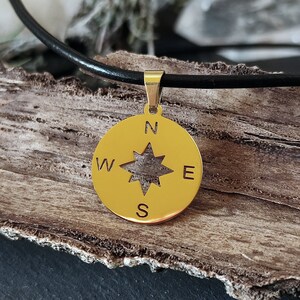 Puede incluir: Collar con colgante de brújula dorado con cordón de cuero negro. El colgante presenta un diseño de rosa de los vientos con los puntos cardinales. El collar se exhibe sobre un trozo de madera.