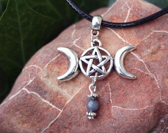 May include: A silver-toned pendant necklace featuring a triple moon design with a central pentagram. The pendant hangs from a black cord and includes a small, gray bead. The necklace is displayed on a reddish-brown stone against a backdrop of green leaves.
