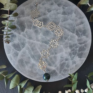 May include: Silver-toned chakra pendant necklace with seven symbolic designs and a dark blue-green gemstone at the bottom. The necklace is displayed on a white, circular surface with green foliage and small white flowers in the background.