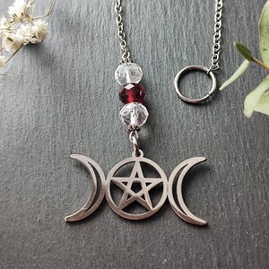 May include: A silver charm with a triple moon and pentagram design, hanging from a silver chain with clear and red crystal beads.