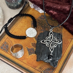 Può includere: Una collezione di gioielli mistici tra cui un ciondolo con nodo celtico argentato su cordino nero, un braccialetto macramè nero con un ciondolo argentato e un anello argentato. Gli oggetti poggiano su un libro vintage con sfondo in velluto rosso scuro.