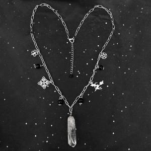 Può includere: Una collana a catena d'argento con un ciondolo di cristallo di quarzo trasparente. La collana ha diversi ciondoli, tra cui un fiore, una stella, un nodo celtico e una fata.