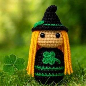 May include: A handmade crochet doll with a black witch hat and dress, adorned with a green shamrock. The doll has yellow yarn hair and button eyes. A green shamrock is in the foreground, set against a blurred green background.