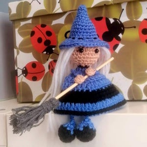 May include: A crocheted witch doll with a blue hat, dress, and shoes. The doll is holding a broom and has long white hair.