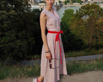 May include: A woman wearing a light pink sleeveless linen dress with a red ribbon tied at the waist. She is standing on a path with a city skyline in the background.