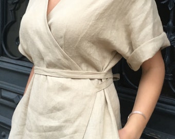 May include: A beige linen wrap dress with a V-neckline and short sleeves. The dress has a tie belt at the waist and pockets on the sides.