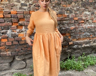 Puede incluir: Un vestido de lino de color melocotón con cuello redondo y mangas 3/4. El vestido tiene una cintura fruncida y una longitud midi.