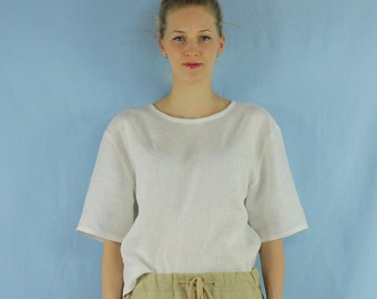 May include: A white linen top with short sleeves and a round neckline. The top is worn with beige pants.