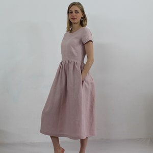 K&ouml;nnte beinhalten: Ein hellrosa Leinenkleid mit Rundhalsausschnitt und kurzen &Auml;rmeln. Das Kleid hat einen gerafften Rock und zwei Taschen.