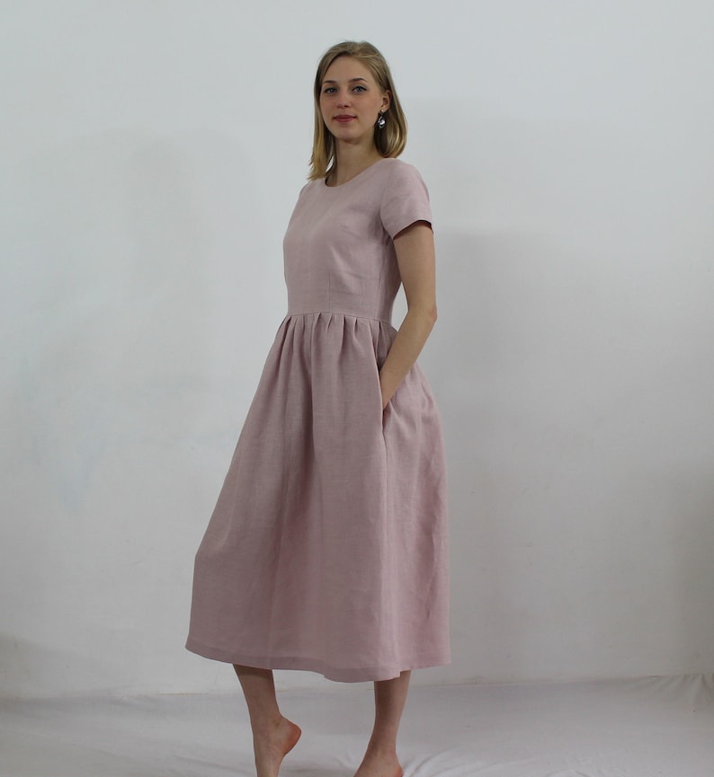 K&ouml;nnte beinhalten: Ein hellrosa Leinenkleid mit Rundhalsausschnitt und kurzen &Auml;rmeln. Das Kleid hat einen gerafften Rock und zwei Taschen.