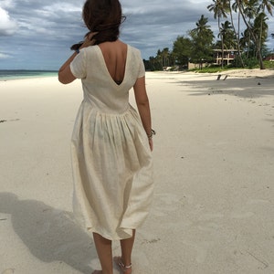 Puede incluir: Una mujer con un vestido de lino blanco con un profundo escote en V y una falda plisada, de pie en una playa de arena blanca.