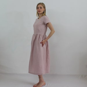 K&ouml;nnte beinhalten: Ein hellrosa Leinenkleid mit Rundhalsausschnitt und kurzen &Auml;rmeln. Das Kleid hat eine geraffte Taille und eine Midi-L&auml;nge.