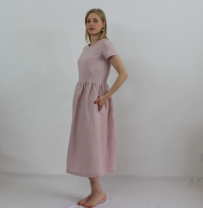 K&ouml;nnte beinhalten: Ein hellrosa Leinenkleid mit Rundhalsausschnitt und kurzen &Auml;rmeln. Das Kleid hat eine geraffte Taille und eine Midi-L&auml;nge.
