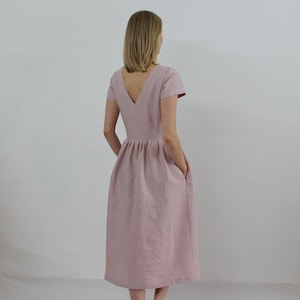 K&ouml;nnte beinhalten: Ein staubiges rosafarbenes Leinenkleid mit V-Ausschnitt und kurzen &Auml;rmeln. Das Kleid hat einen gerafften Rock und Taschen.