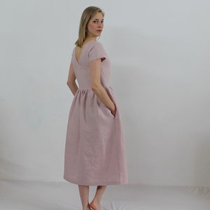 K&ouml;nnte beinhalten: Ein hellrosa Leinenkleid mit V-Ausschnitt, kurzen &Auml;rmeln und einem gerafften Rock. Das Kleid hat Taschen an den Seiten.