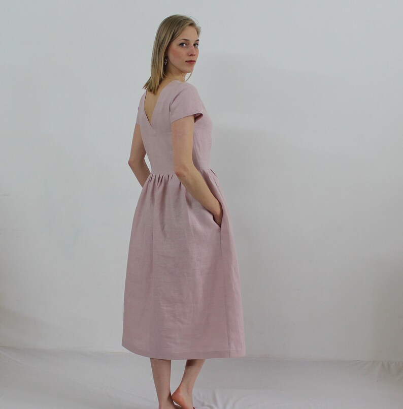K&ouml;nnte beinhalten: Ein hellrosa Leinenkleid mit V-Ausschnitt, kurzen &Auml;rmeln und einem gerafften Rock. Das Kleid hat Taschen an den Seiten.