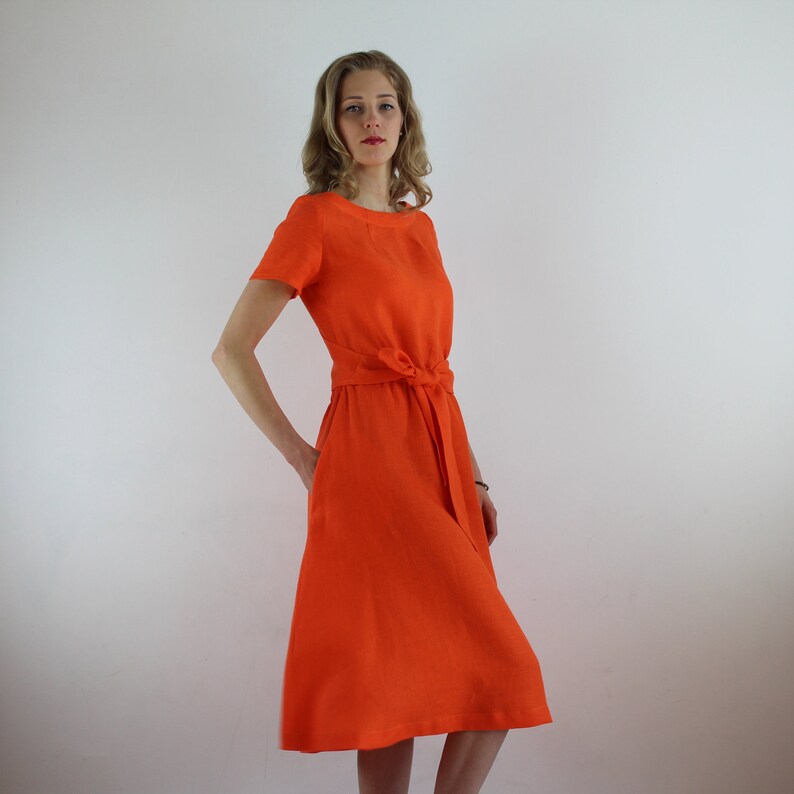 May include: A bright orange linen dress with a tie belt at the waist. The dress has short sleeves and a scoop neckline.