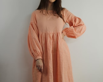 Puede incluir: Un vestido de lino de color melocotón con mangas largas y falda fruncida. El vestido tiene un corte holgado y un escote redondo.