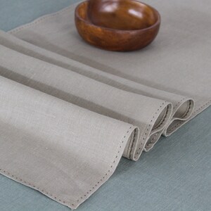 May include: A beige linen table runner with a stitched edge, folded in thirds, on a blue surface. A wooden bowl is in the background.