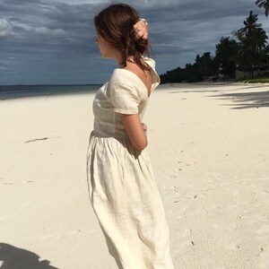 Puede incluir: Una mujer con un vestido de lino blanco se encuentra en una playa de arena blanca. El vestido tiene un escote redondo y mangas cortas. La mujer mira hacia el oc&eacute;ano.