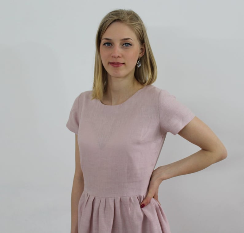 K&ouml;nnte beinhalten: Eine Frau tr&auml;gt ein hellrosa Leinenkleid mit kurzen &Auml;rmeln und einem runden Ausschnitt. Das Kleid hat einen gerafften Rock.