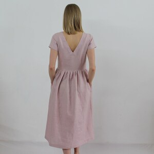 K&ouml;nnte beinhalten: Ein staubiges rosafarbenes Leinenkleid mit V-Ausschnitt und kurzen &Auml;rmeln. Das Kleid hat einen gerafften Rock und Taschen.