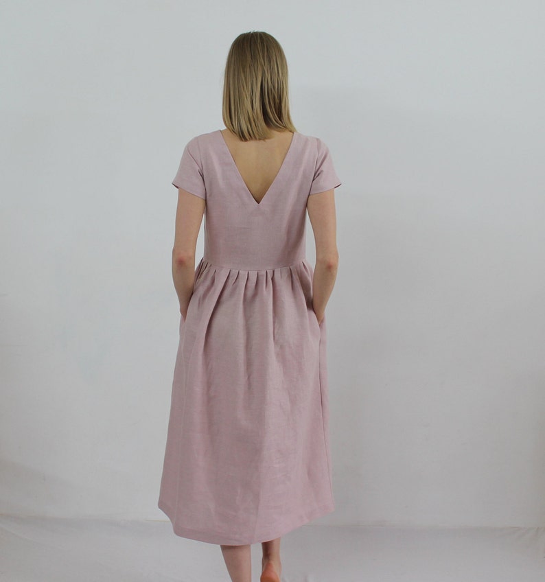 K&ouml;nnte beinhalten: Ein staubiges rosafarbenes Leinenkleid mit V-Ausschnitt und kurzen &Auml;rmeln. Das Kleid hat einen gerafften Rock und Taschen.