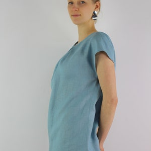 May include: A woman wearing a light blue linen top with short sleeves and gray pants. She is standing with her arms crossed and looking at the camera.