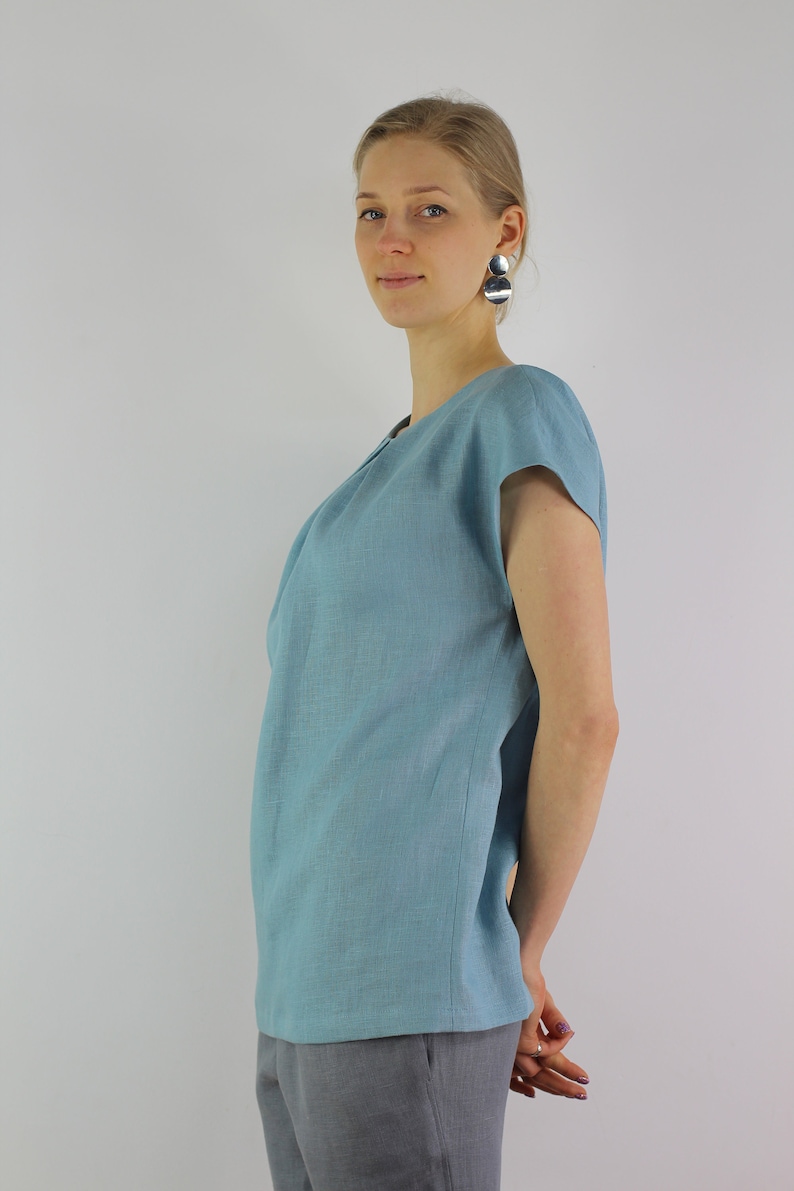 May include: A woman wearing a light blue linen top with short sleeves and gray pants. She is standing with her arms crossed and looking at the camera.