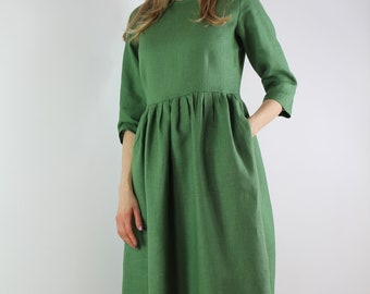 Puede incluir: Un vestido de lino verde con mangas tres cuartos y cintura fruncida. El vestido presenta un cuello redondo y bolsillos. El vestido es usado por una modelo y llega a la rodilla.
