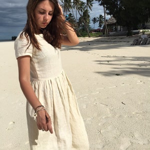Puede incluir: Una mujer con un vestido de lino blanco con falda fruncida y mangas cortas. Est&aacute; de pie en una playa de arena blanca con palmeras en el fondo.