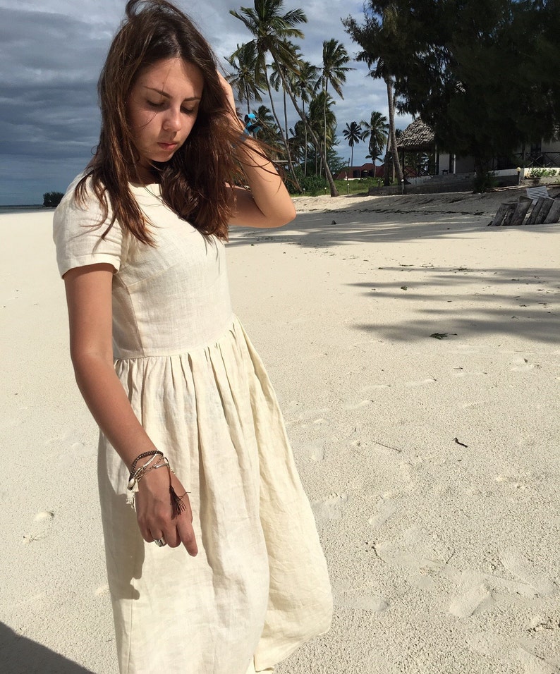 Puede incluir: Una mujer con un vestido de lino blanco con falda fruncida y mangas cortas. Est&aacute; de pie en una playa de arena blanca con palmeras en el fondo.