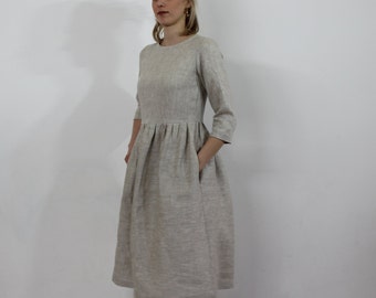 Puede incluir: Un vestido de lino beige claro con cuello redondo y mangas 3/4. El vestido tiene una falda fruncida y bolsillos a los lados.