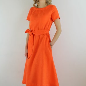 May include: A bright orange linen dress with a short sleeve and a tie belt. The dress has a simple, classic design.