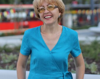May include: A woman with short blonde hair is wearing a bright blue wrap dress and smiling. She is wearing glasses with yellow lenses.