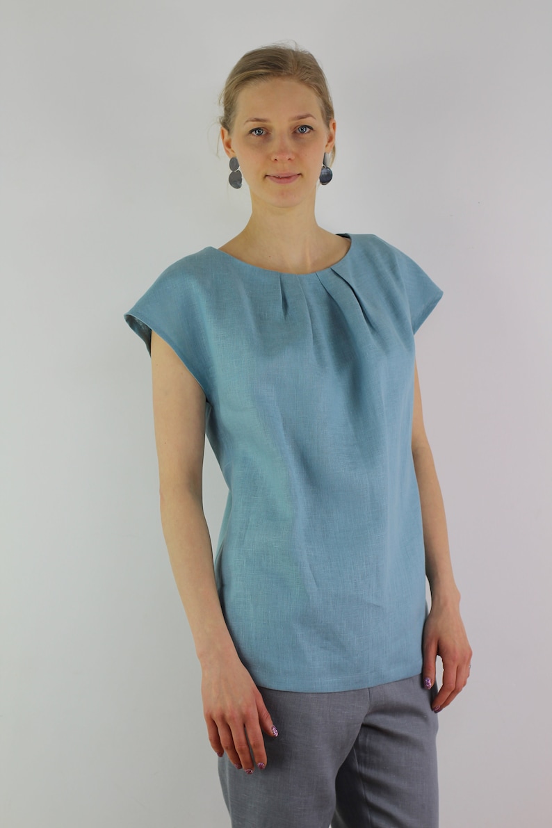 May include: A light blue linen top with a pleated neckline and short sleeves. The top is loose-fitting and has a relaxed silhouette.