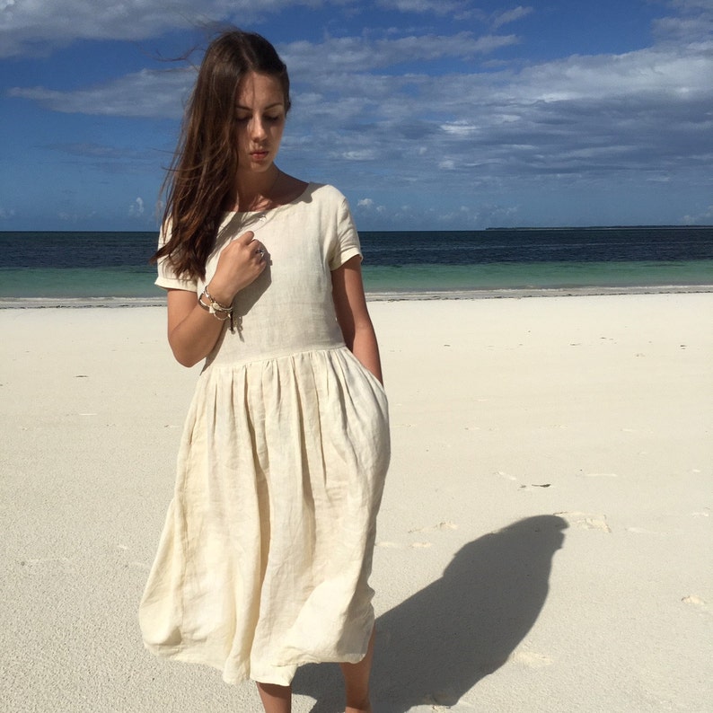 Puede incluir: Una mujer con un vestido de lino blanco se encuentra en una playa de arena blanca con un oc&eacute;ano azul de fondo.