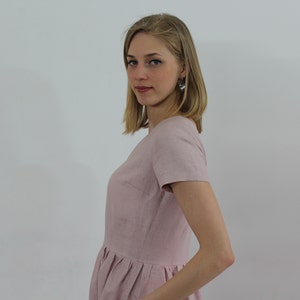 K&ouml;nnte beinhalten: Ein hellrosa Leinenkleid mit Rundhalsausschnitt und kurzen &Auml;rmeln. Das Kleid hat einen gerafften Rock und einen taillierten Oberteil.
