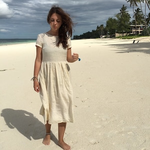 Puede incluir: Una mujer con un vestido de lino blanco camina por una playa de arena blanca. El vestido tiene mangas cortas y una falda plisada.