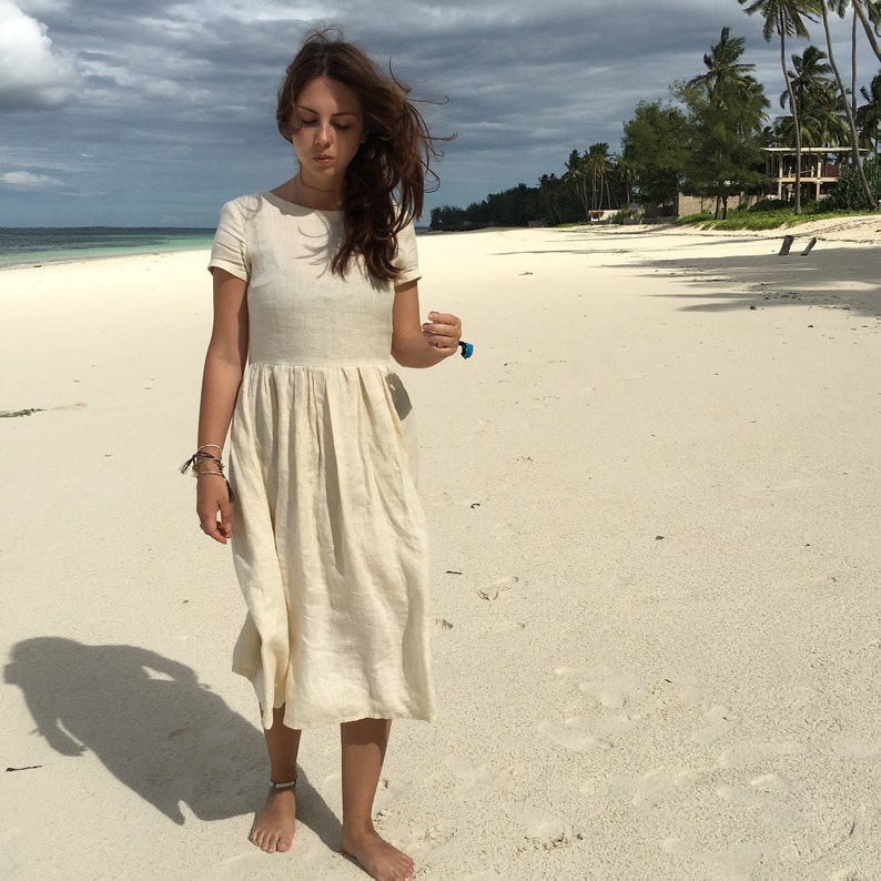 Puede incluir: Una mujer con un vestido de lino blanco camina por una playa de arena blanca. El vestido tiene mangas cortas y una falda plisada.